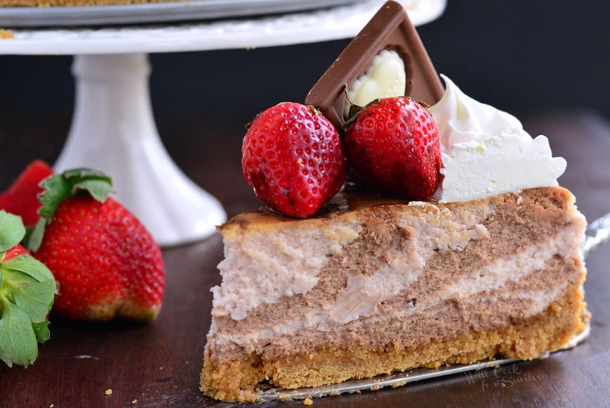 Chocolate Strawberry Marbled Cheesecake on a table with strawberries around it and a cake stand in the background