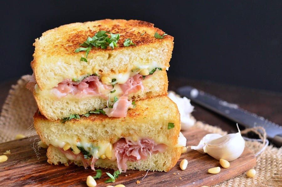 two halves of Italian grilled cheese with prosciutto stacked on the cutting board with garlic cloves around.