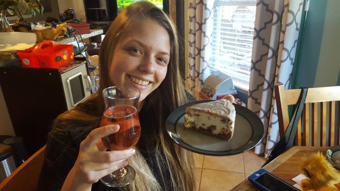 my sister holding a plate with a piece of cheesecake and in one hand and a drink in the other
