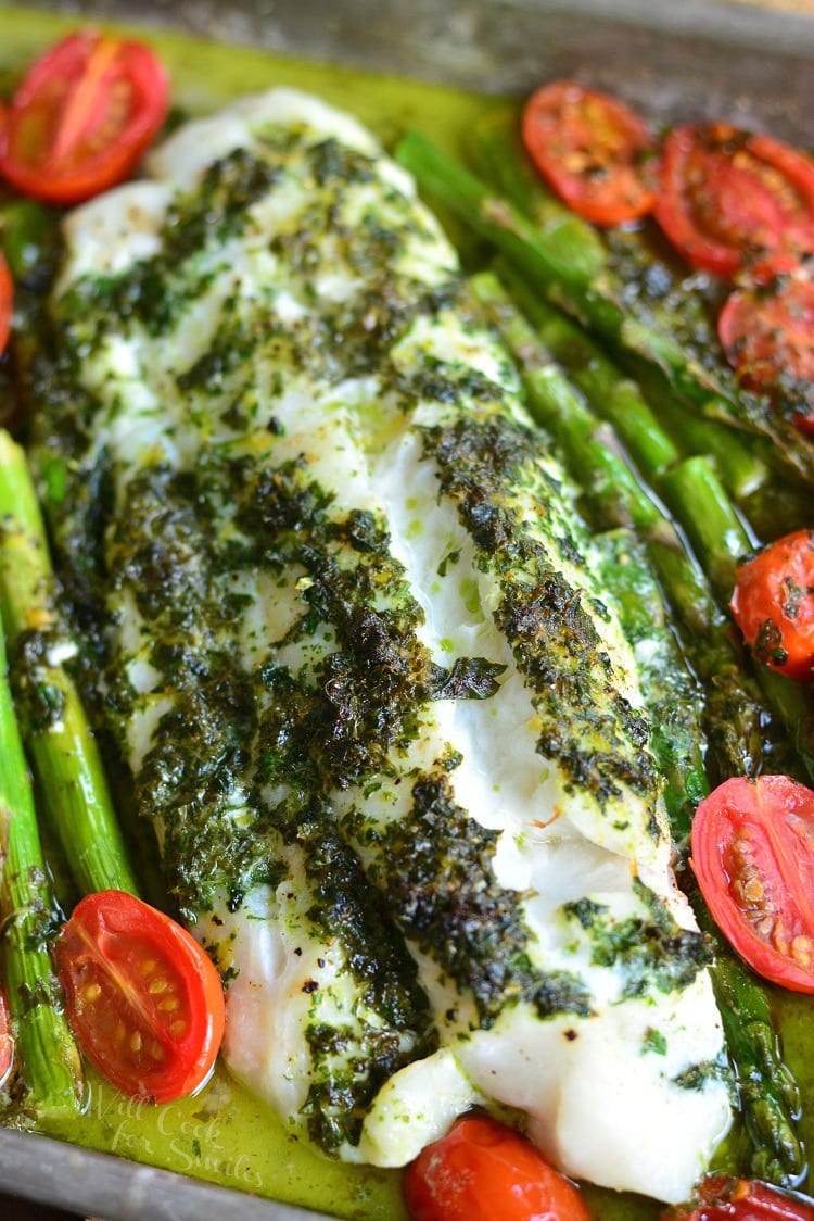 a large baked cod filet covered in herbs on the baking pan with asparagus and cherry tomatoes. 