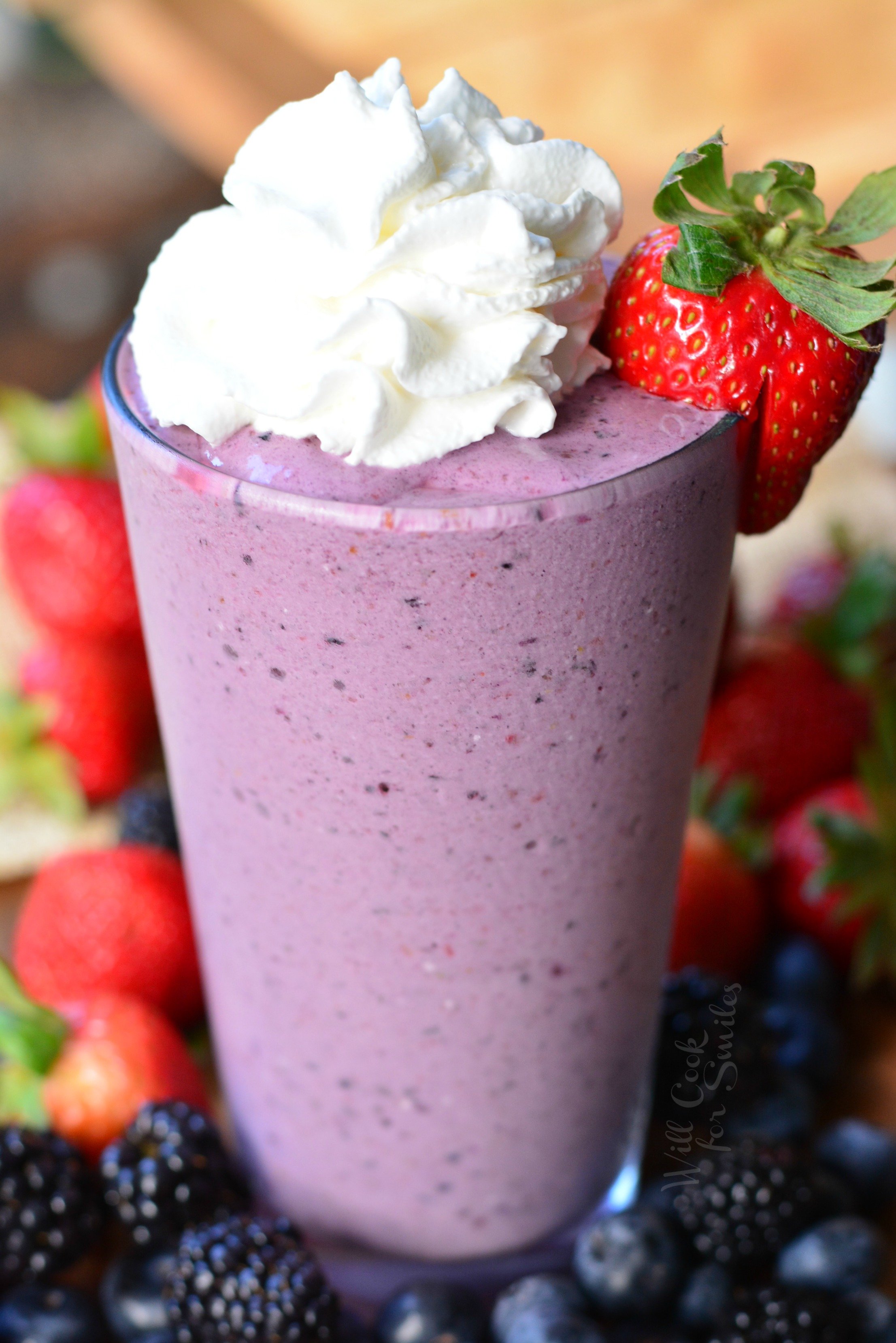 Berry Milkshake in a class with whip cream and strawberry to garnish on a wood table with blackberries, strawberries, and blueberries