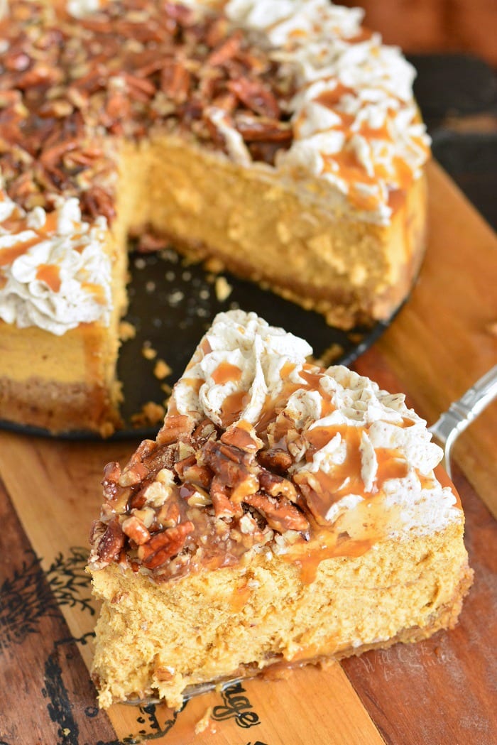 Pumpkin Cheesecake with Pecans and Caramel slice on a wooden cutting board with the rest of the cheesecake in the background