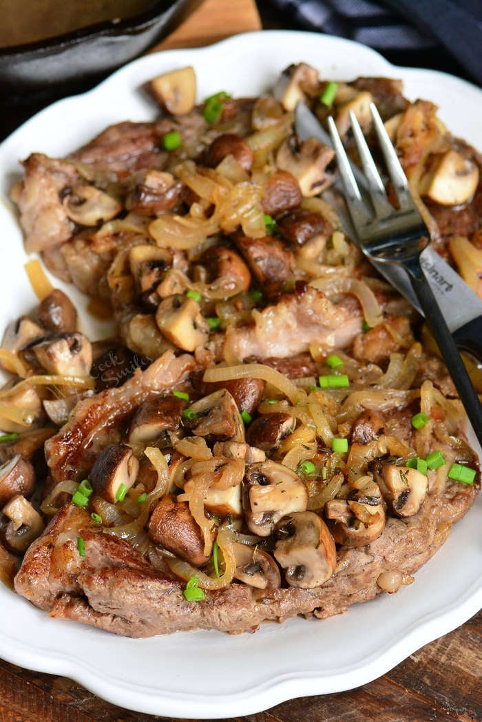 rib eyes steaks on the plate topped with mushrooms and onions with a fork.
