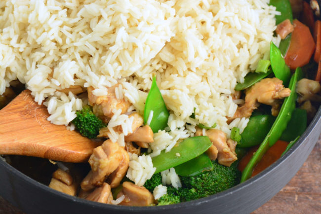 adding rice to chicken and vegetables in a pan with a wooden spoon