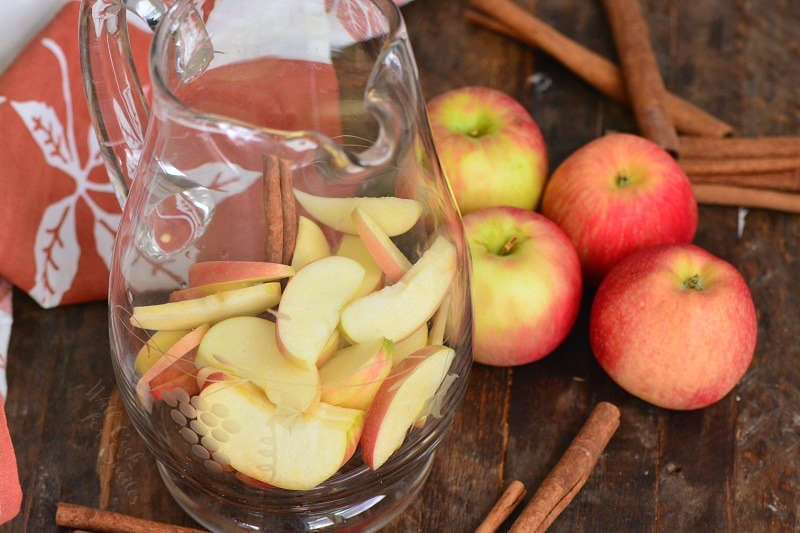 apple in a pitcher sitting on a wood table with apples and cinnamon sticks