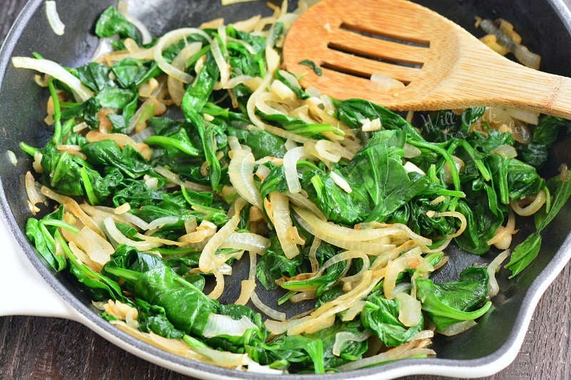 spinach and onions cooking in a cast iron pan with a wooden spatula