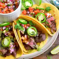 three steak tacos topped with jalapeno rings and pico on a light brown plate with a bowl of pico next to them and some lime slices around