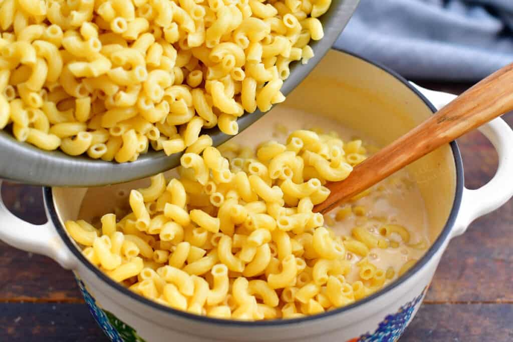 cooked pasta being poured into a pot of cheese sauce