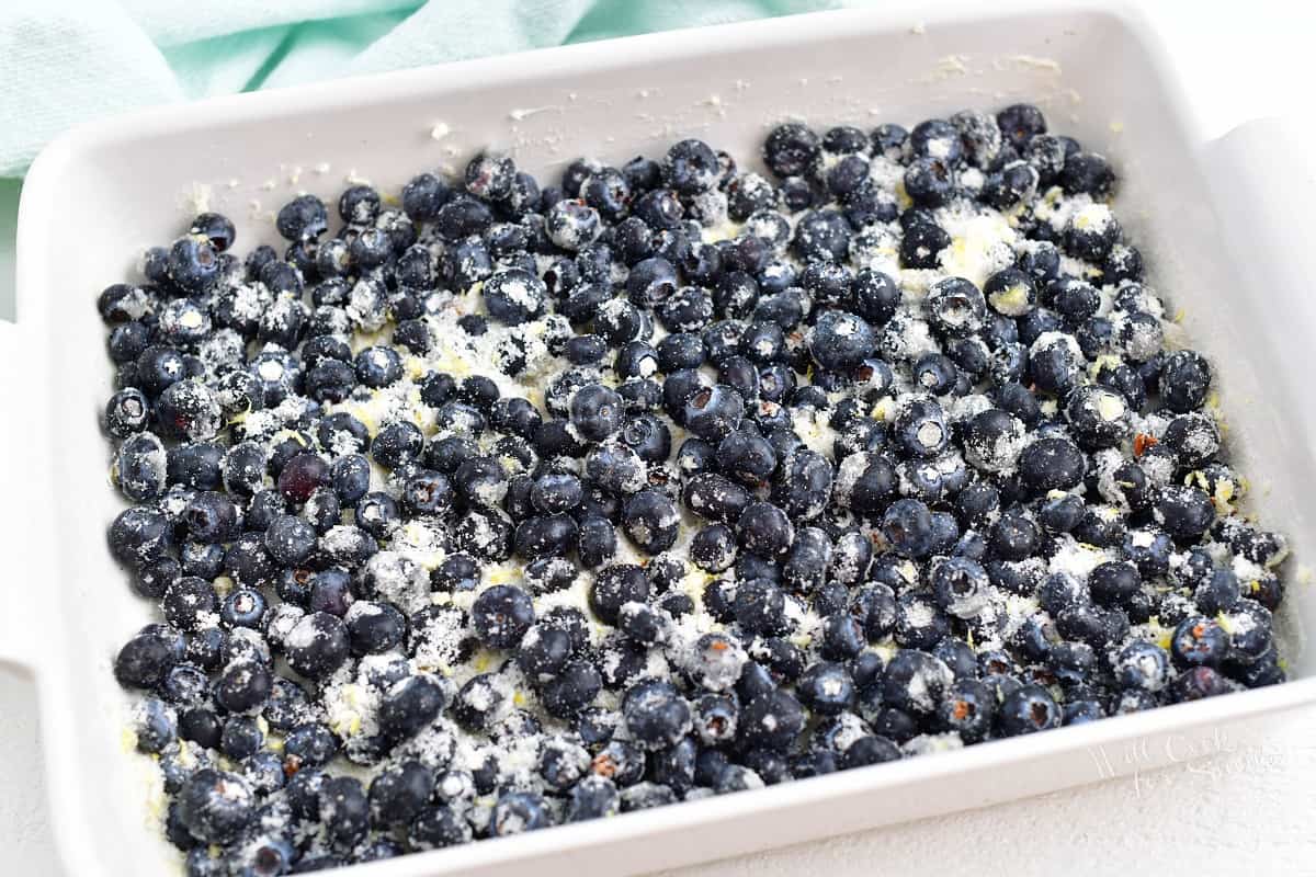 Blueberries and sugar are mixed together and spread evenly in a large white casserole dish.