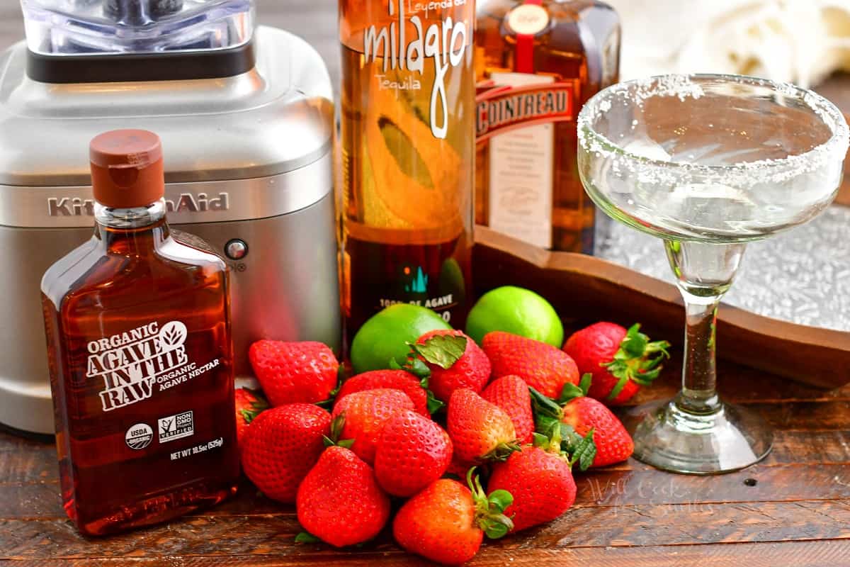 The ingredients for strawberry margaritas are laid out on a wooden surface.