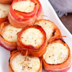 Scallops wrapped in bacon are piled on a rectangular white plate.