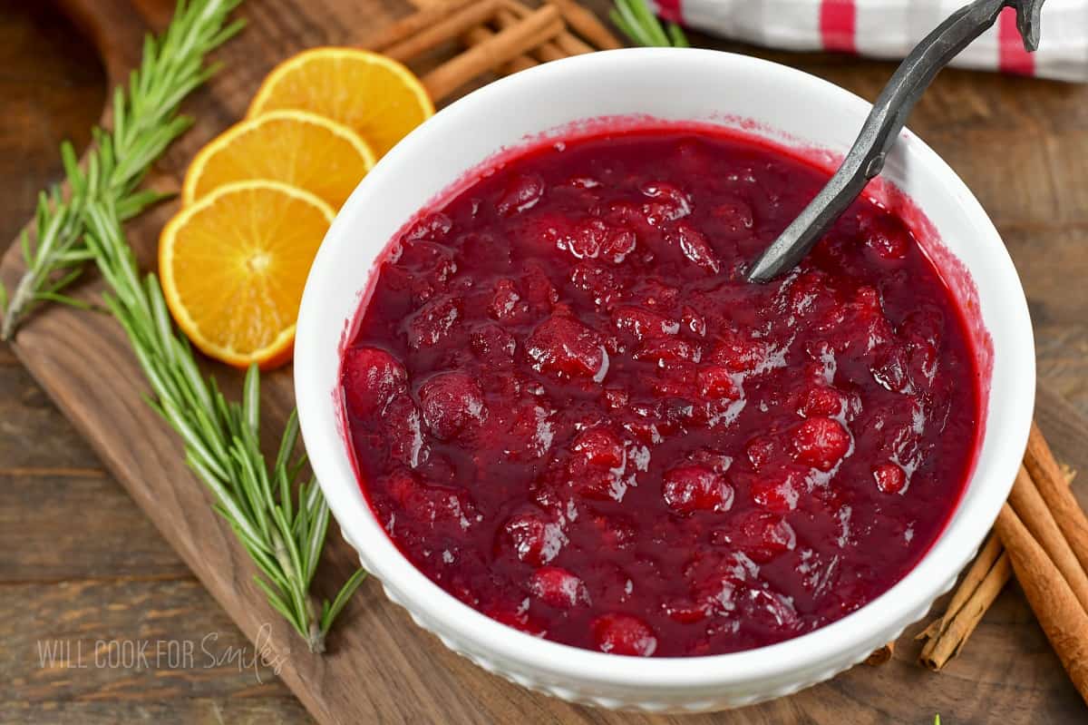 cranberry sauce in a white bowl with spoon, orange slices, and rosemary.