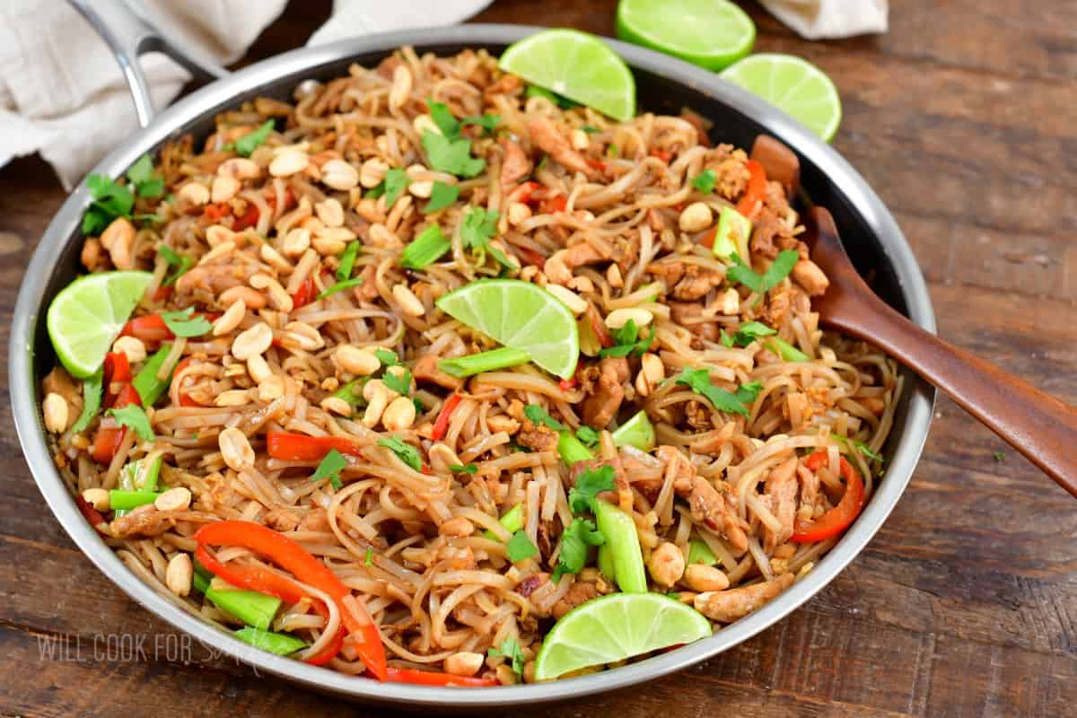 A wooden spoon is placed in a skillet filled with garnished pad Thai.