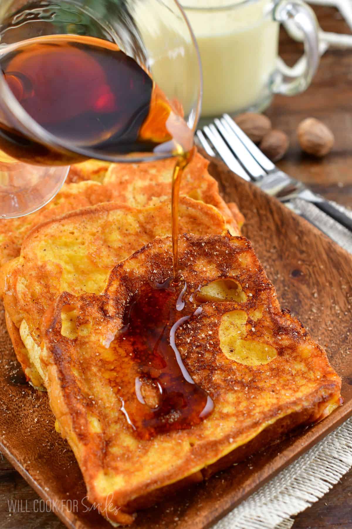 pouring amber maple syrup over Eggnog French Toast slices stacked on a plate.