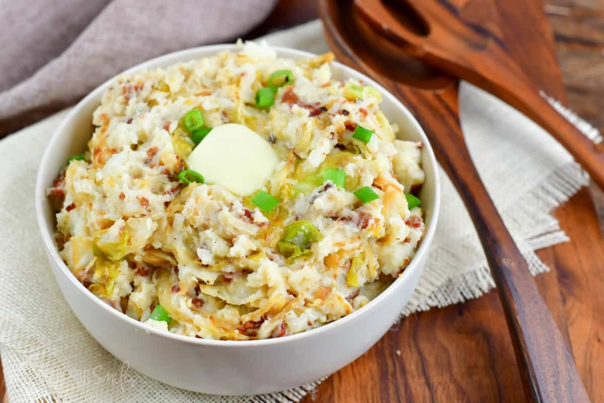 colcannon mashed potaotes in a bowl with a slice of butter and green onions on top as garnish and a set of wooden serving spoons to the left.