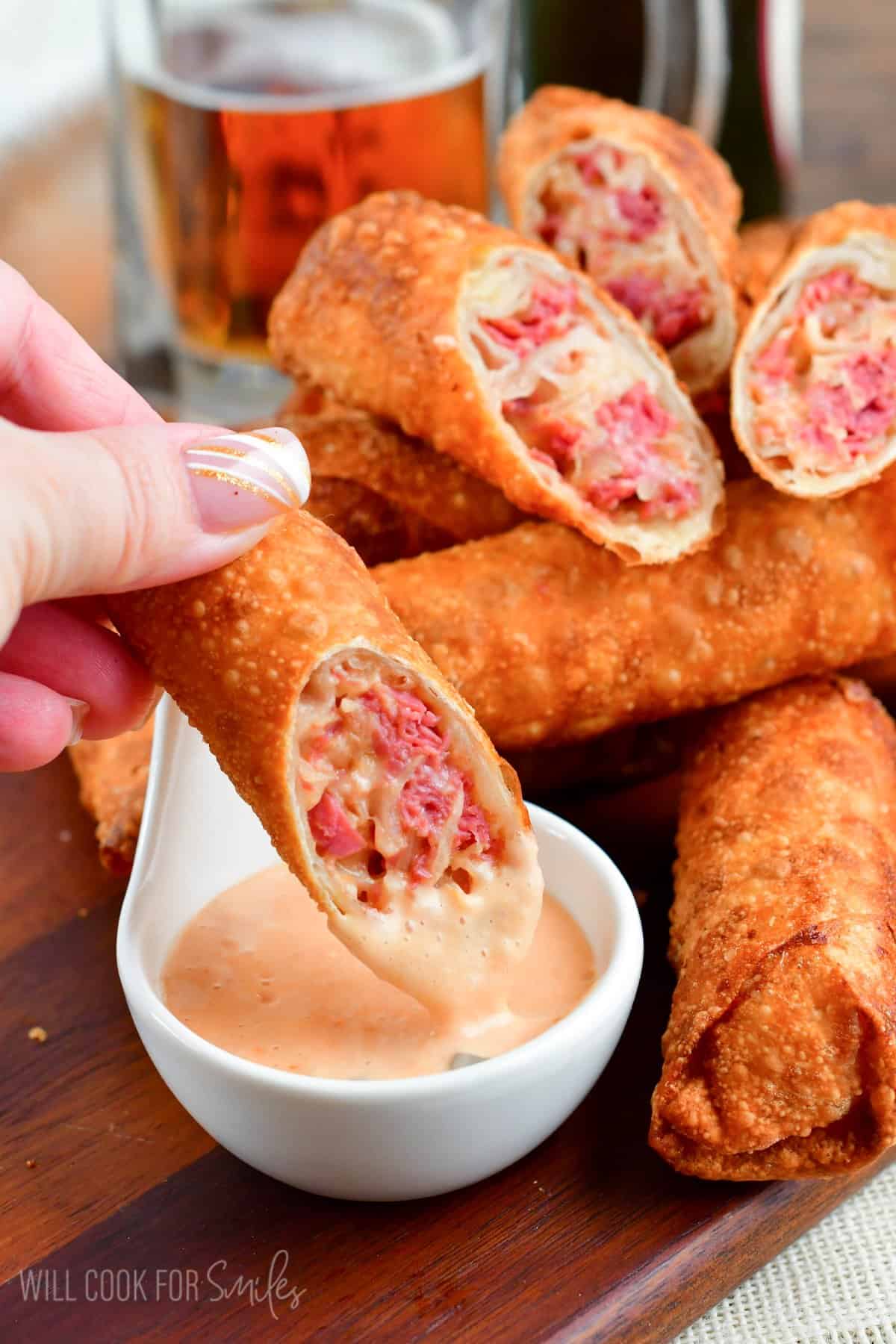 Dipping a reuben egg roll into dressing with the rest of the egg rolls on a wooden tray.