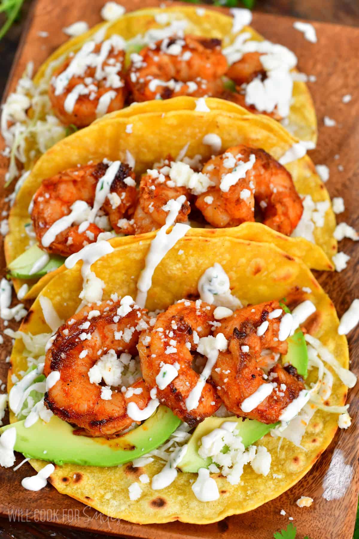 Three shrimp tacos on a wood plate with creme, avocado, an dcheese.