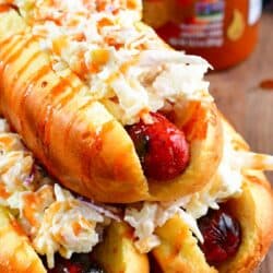 Three Sriracha Honey Hot Dogs with on them stacked on a wood surface with a bottle of Sriracha in the background.