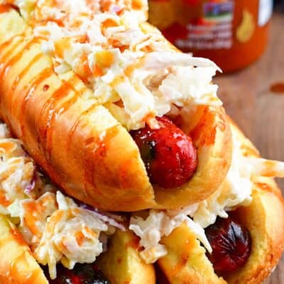 Three Sriracha Honey Hot Dogs with on them stacked on a wood surface with a bottle of Sriracha in the background.