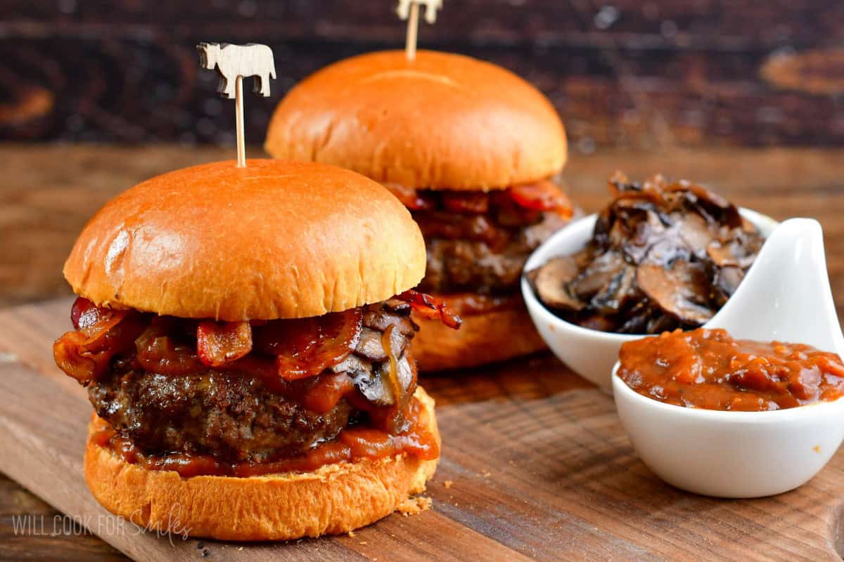 Two burgers on a bun with sauteed mushrooms, bacon, and whiskey sauce with a side of sauce and mushrooms in little bowls.