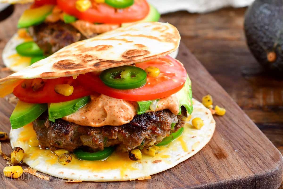 Quesadilla burger with tomato, avocado, and jalapenos on a on a wood cutting board.