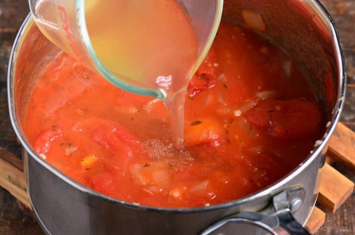 Adding the broth from a measuring cup into the pot of tomato basil soup.
