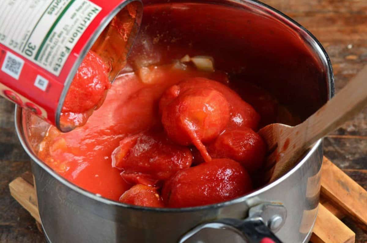 Adding whole tomatoes into a pot with a wooden spoon to the side.