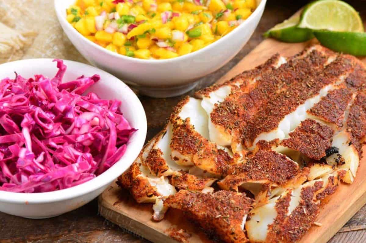 Blackedned tilapia flaked apart on a wood board with cabbage and mango salsa in bowls nex to it.