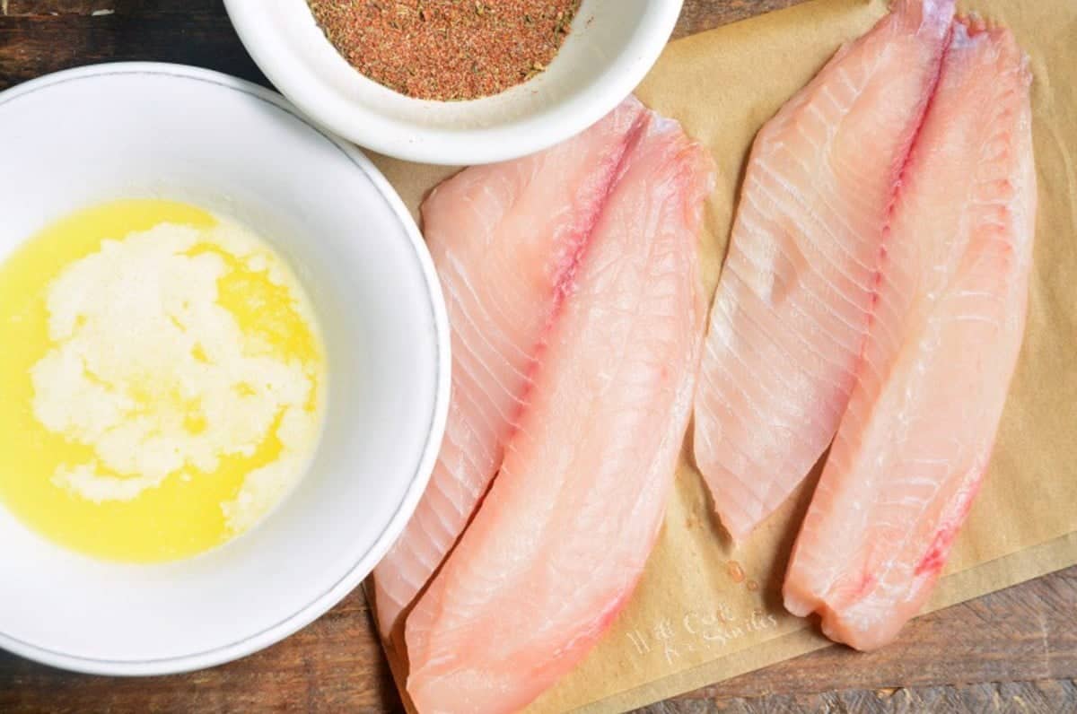 two peices of tilapia on a piece of parchment paper with a bowl of butter and blackening seasoning next to it.