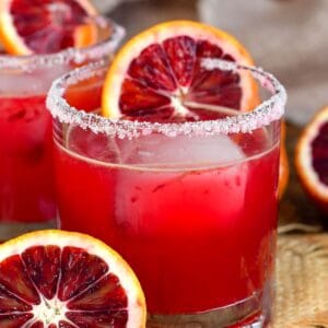 Blod orange margarita in a salt rimmed glass with a slice of orange on the glass as garnish and half an orange next to the glass.
