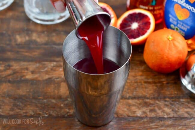 Pouring the blood orange juice into the metal shaker with oranges in the background.