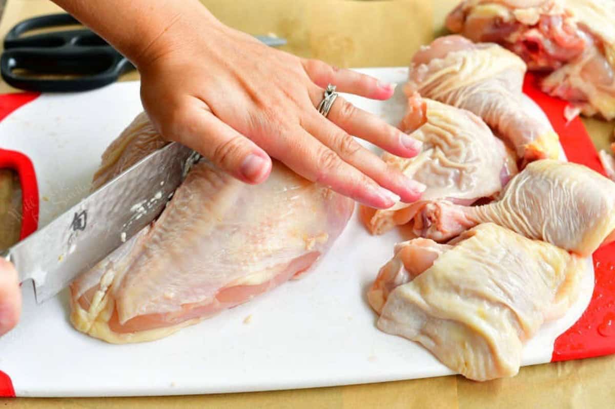 using a large knife to cut the chicken breast in half on a cutting board.