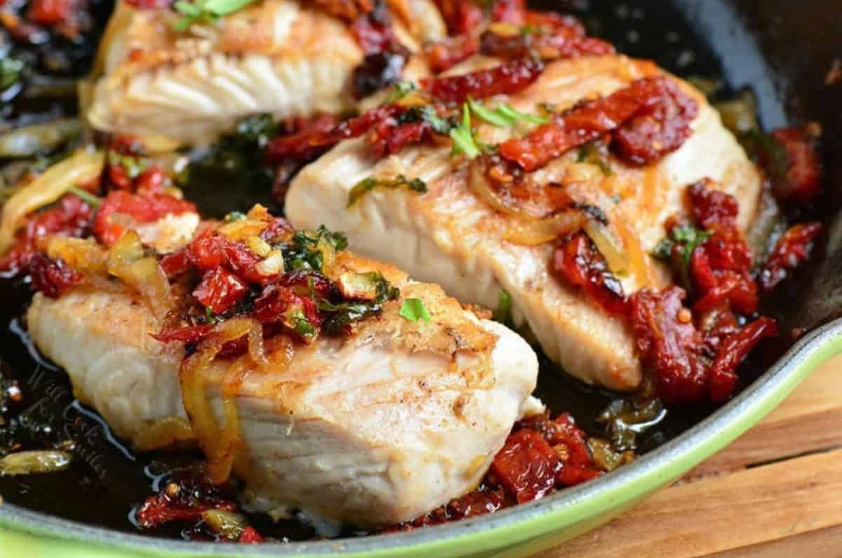 three baked mahi mahi filets in the skillet with sauteed sun dried tomatoes, onion, and basil on top.
