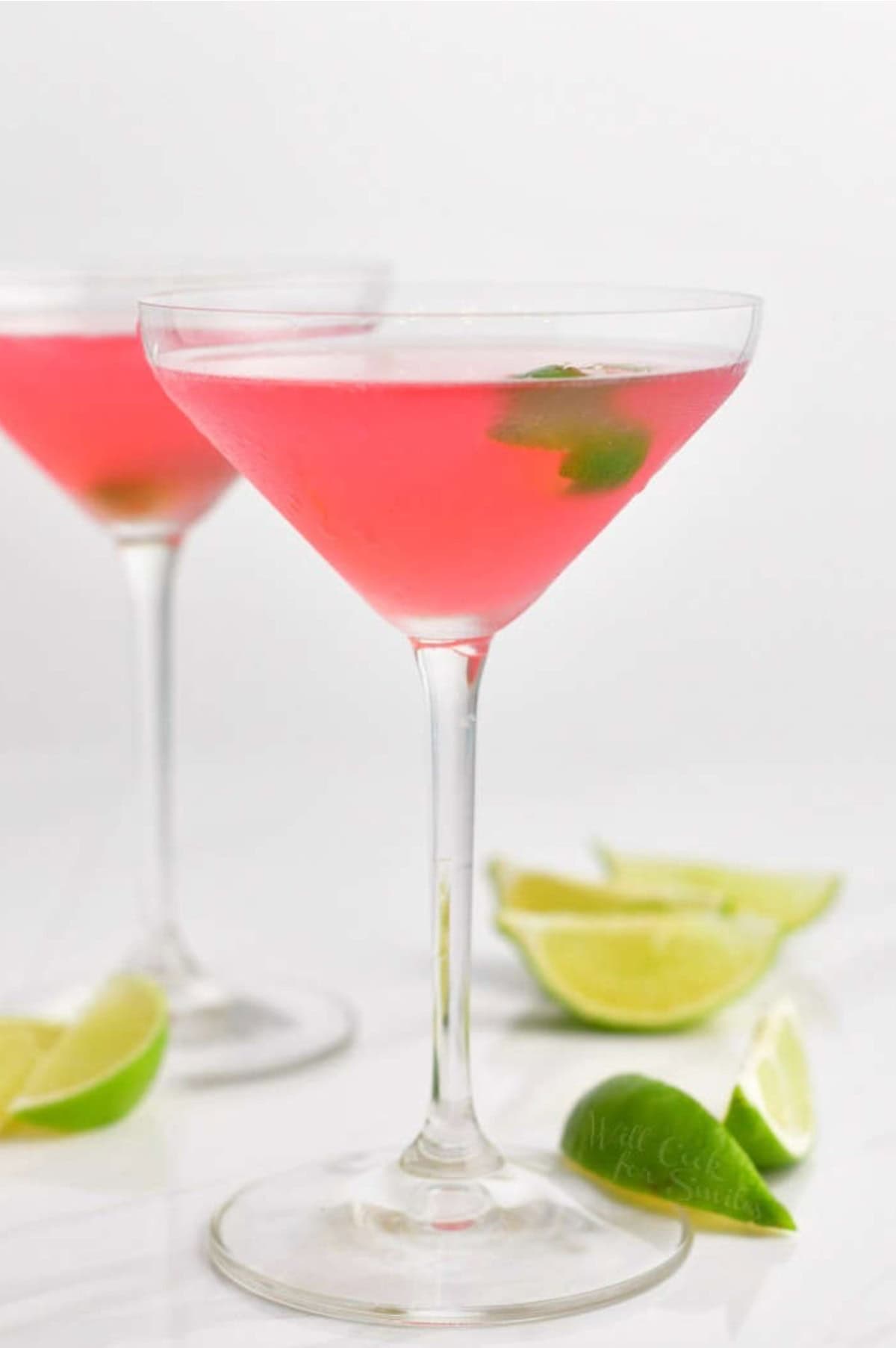 two tall martini glasses filled with cosmopolitan cocktail and lime twist.