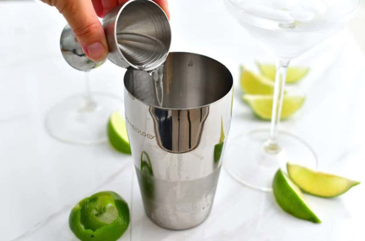 pouring vodka into cocktail shaker next to a martini glass and limes.