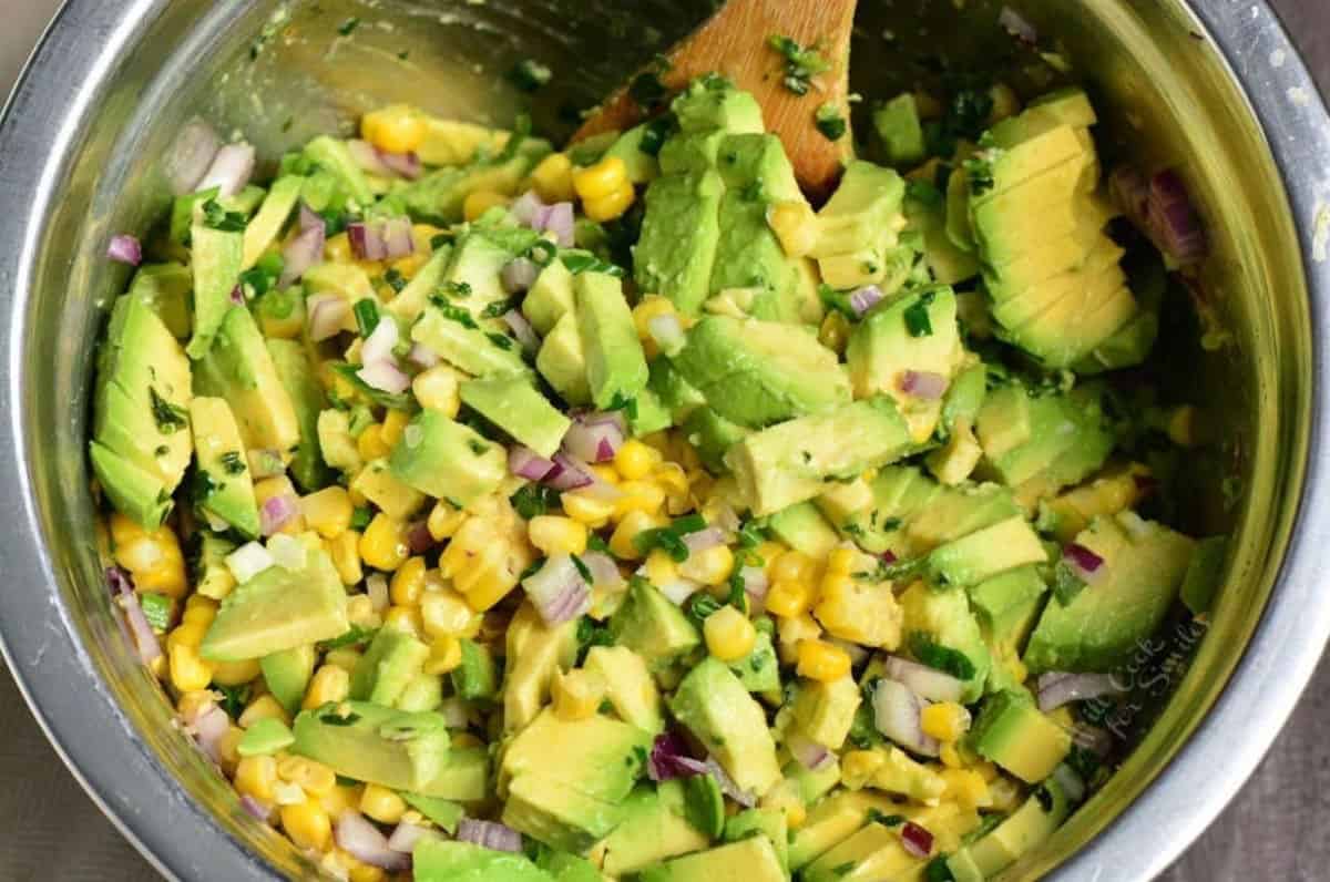 Avocado, corn, and red onion for avocado corn salad in a bowl with a wooden spoon.