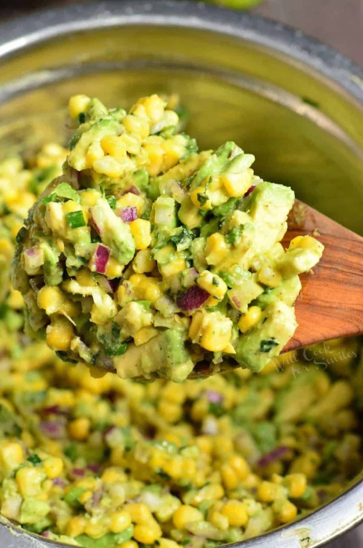 Using a wooden spoon to scoop out some avocado corn salad out of a bowl.