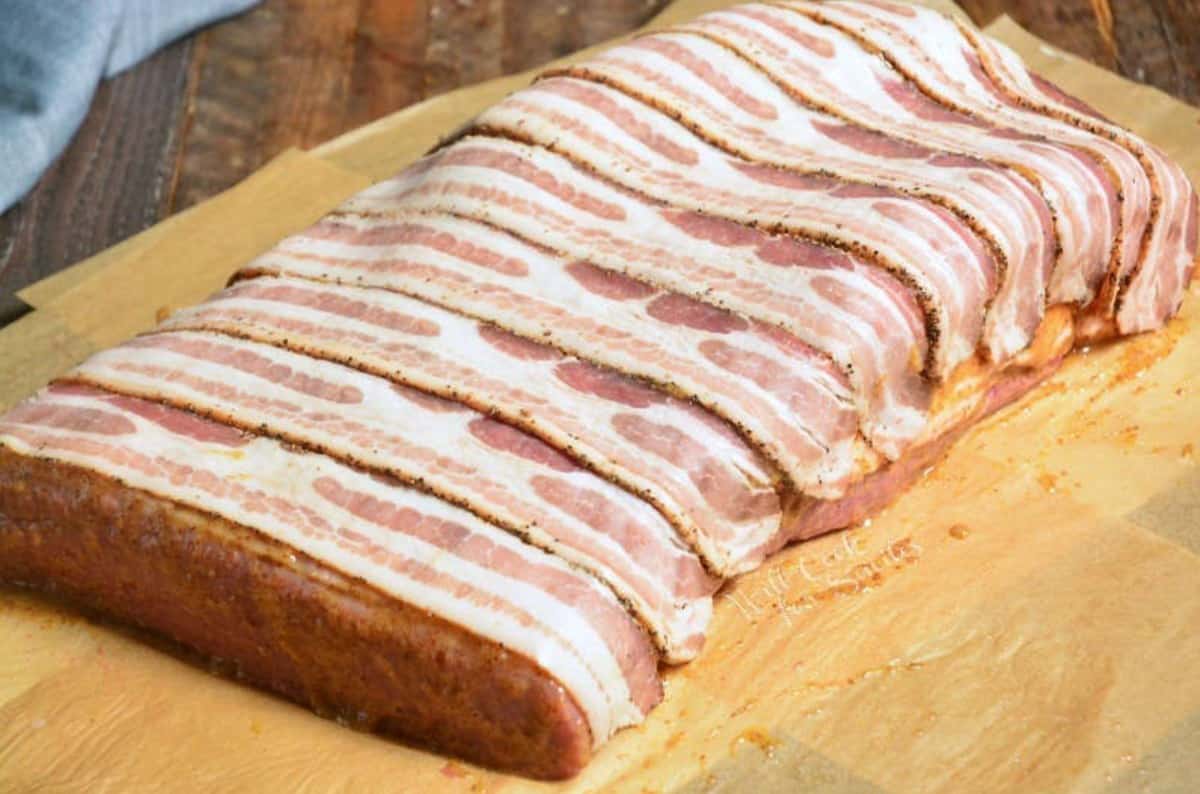 whole brisket wrapped in bacon strips on parchment paper before cooking.