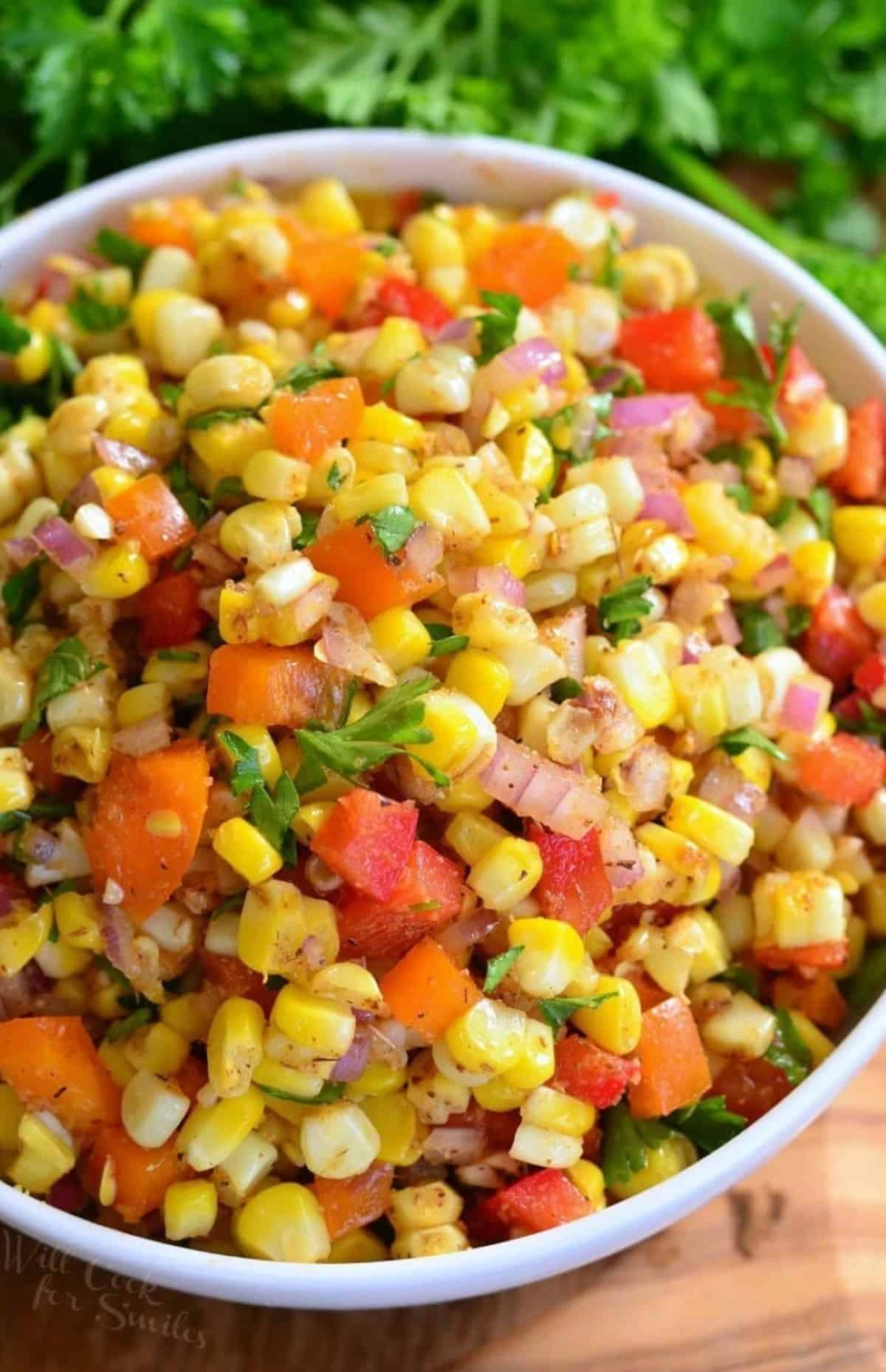 A bowl of Cajun corn salad with herbs to the side on a wood surface.