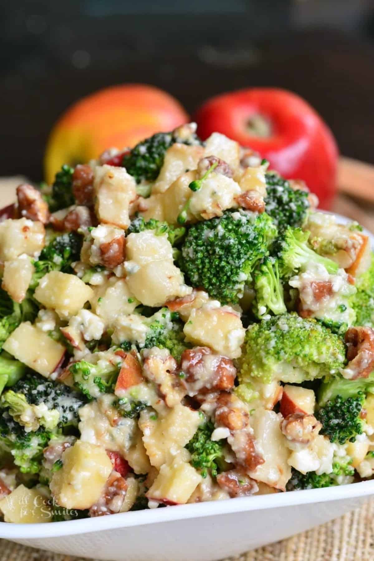 a white bowl full of broccoli salad with apples, nuts, and blue cheese.