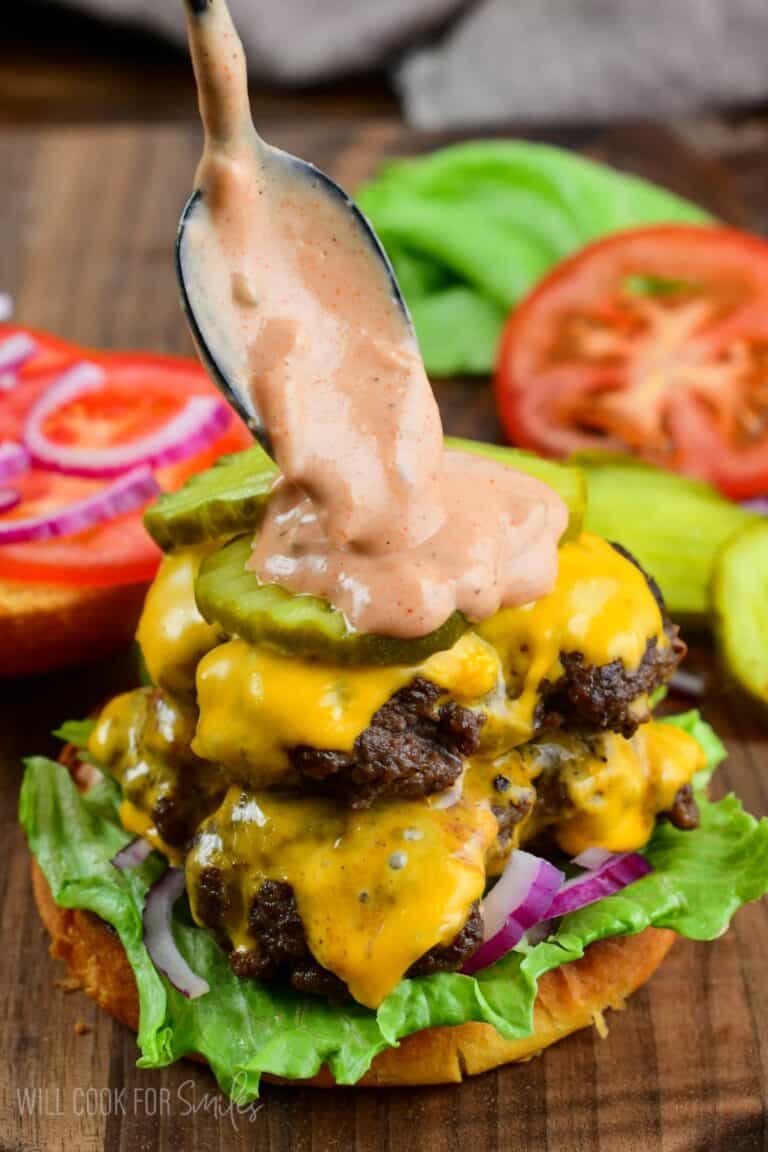 adding creamy burger sauce with a spoon on top of a juicy smashed burger with lettuce, pickle, onion, and cheese.