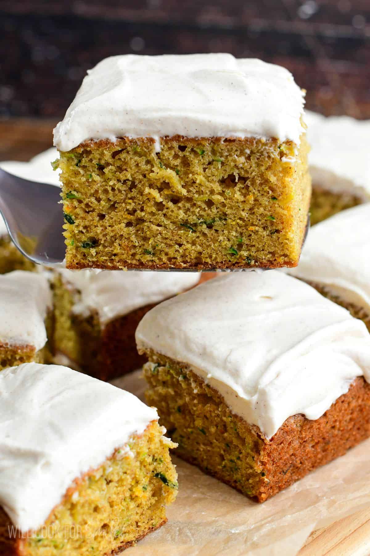 Zucchini cake square on a spatula with the rest of the cake cut into squares on parchment paper.