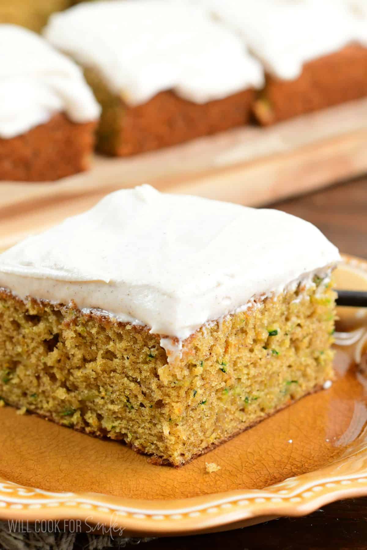 a slice of zucchini cake covered in cream cheese frosting on a plate next to more cake slices.