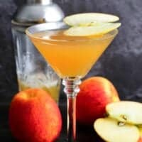 apple martini in a martini glass with a slice of apple on top as garnish with 3 apples one sliced on table.