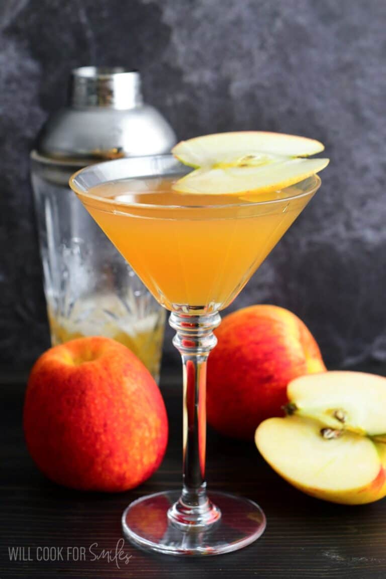 apple martini in a martini glass with a slice of apple on top as garnish with 3 apples one sliced on table.