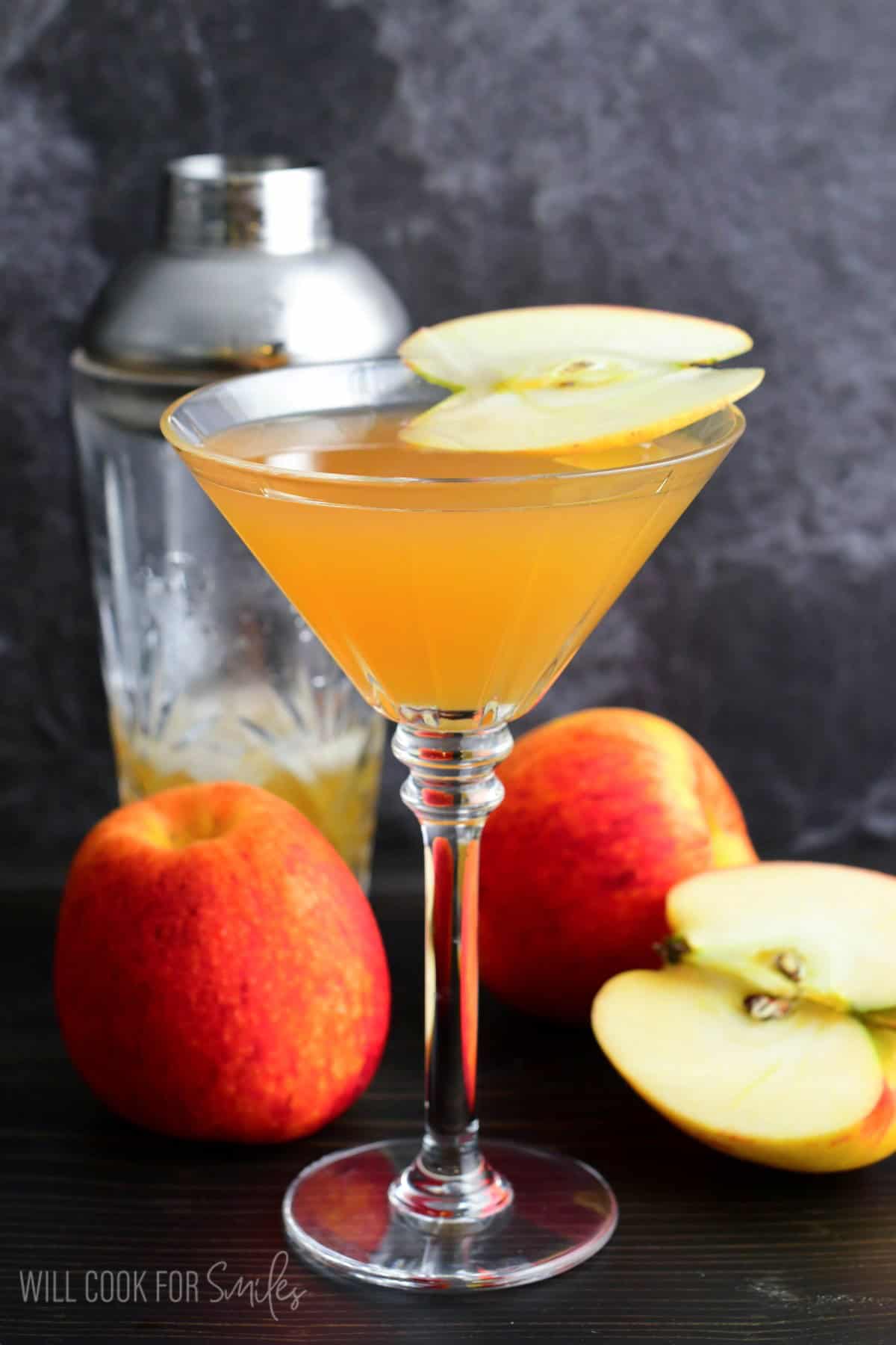 apple martini in a martini glass with a slice of apple on top as garnish with 3 apples one sliced on table.