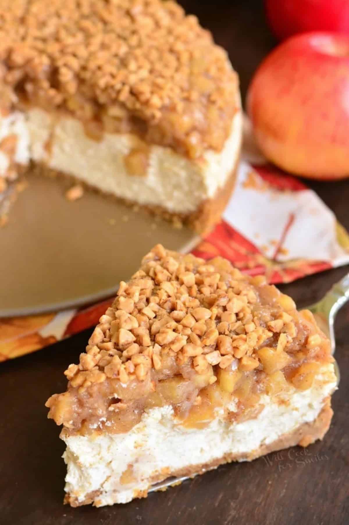 a slice of apple pie cheesecake next to remaining cheesecake and apples.