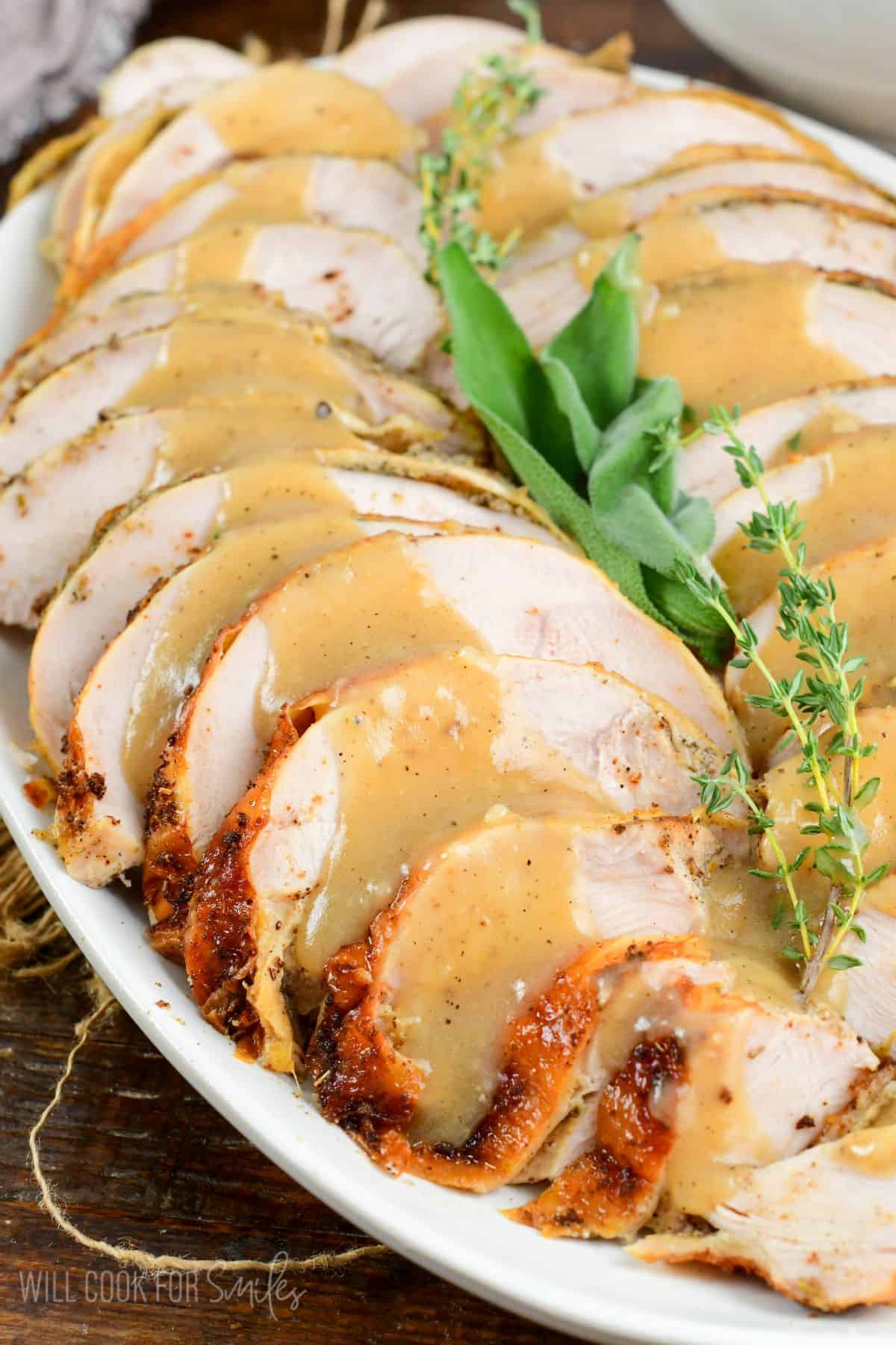 Juicy and moist sliced roasted turkey breast on a plater with pan drippings gravy on top, sage and thyme in the middle as garnish.