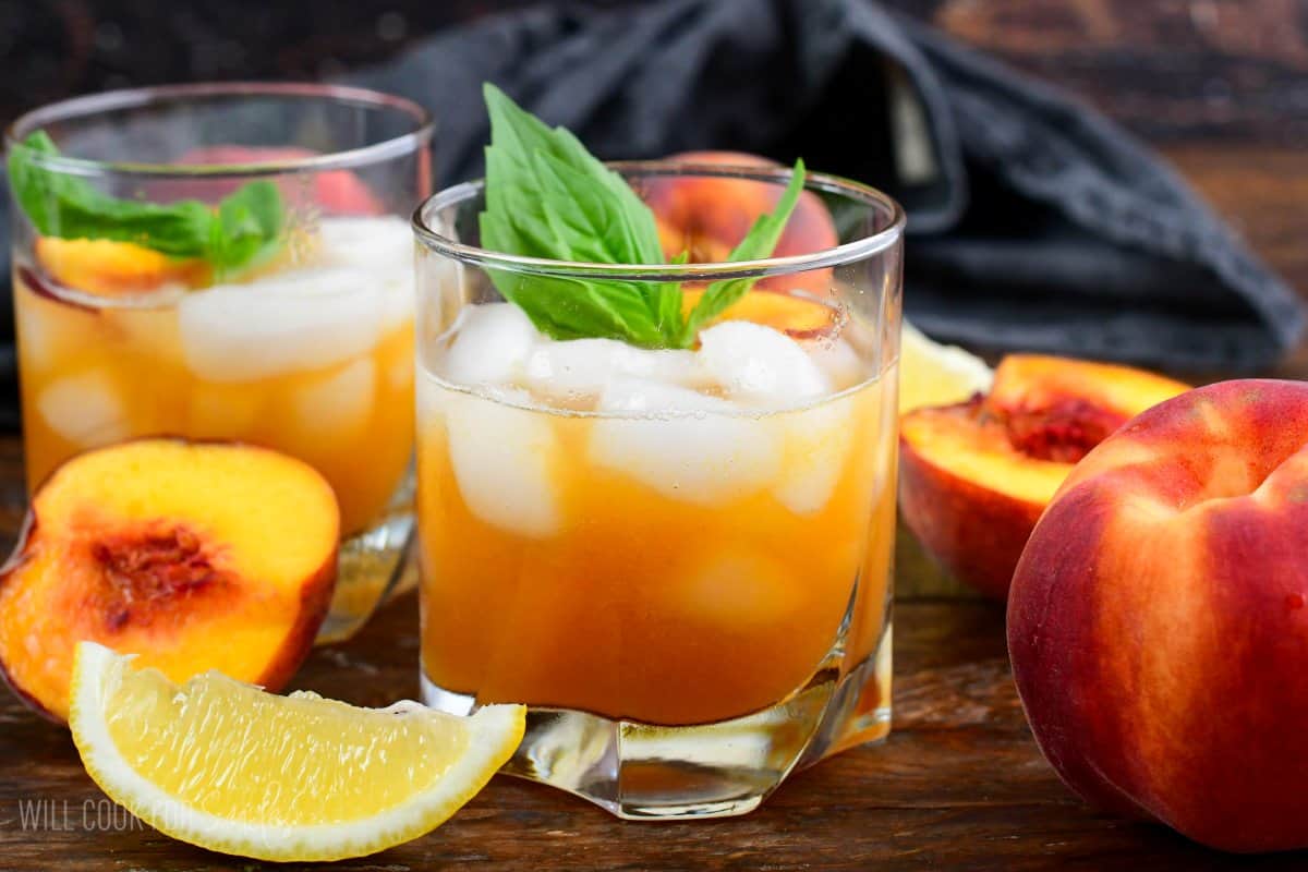 two glasses of peach smash bourbon in a glass with ice and peaches and mint on top as garnish on a wood surface.