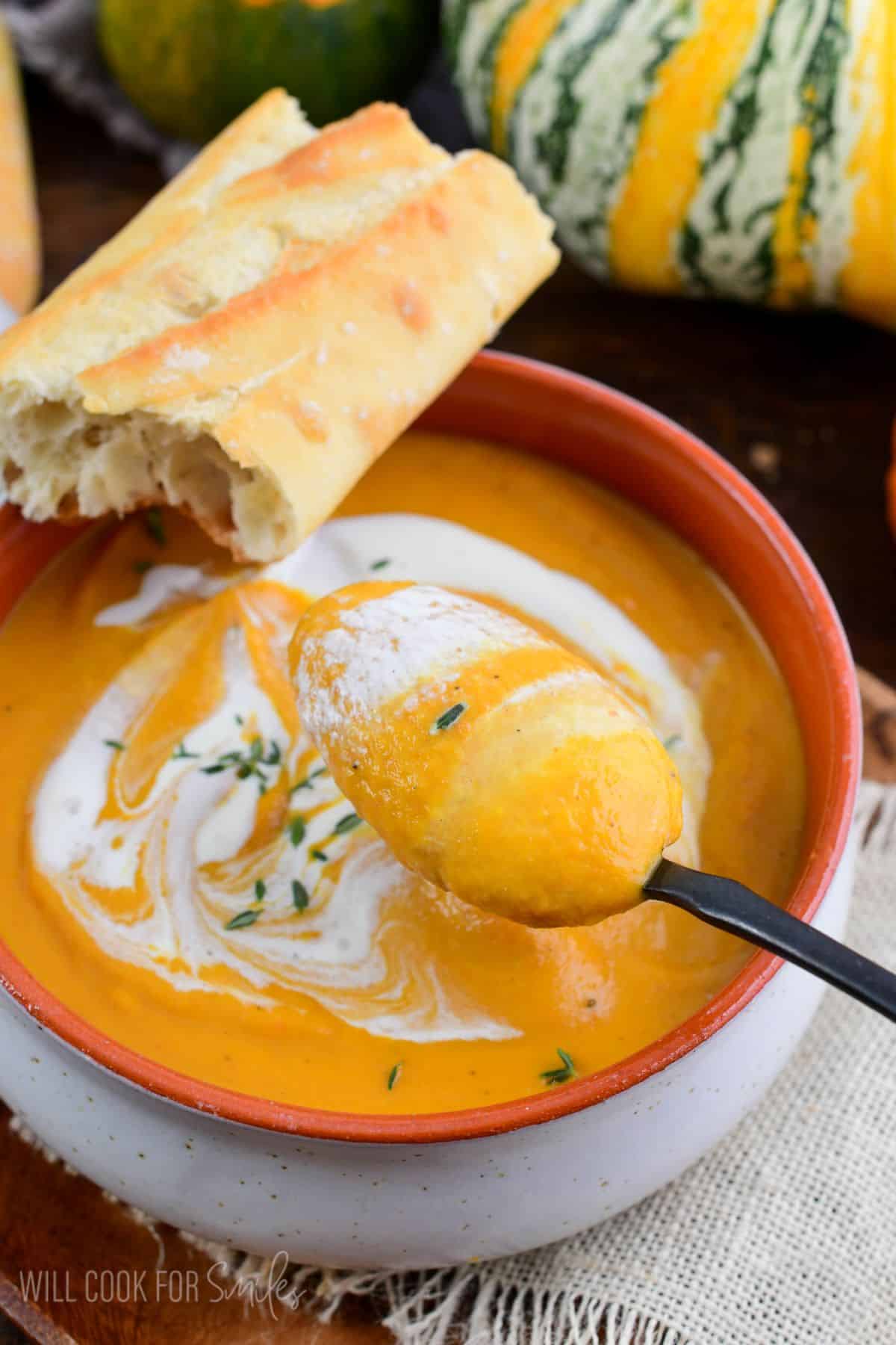 pumpkin soup in a bowl with a spoon scooping some out and bread across the back of the bowl.
